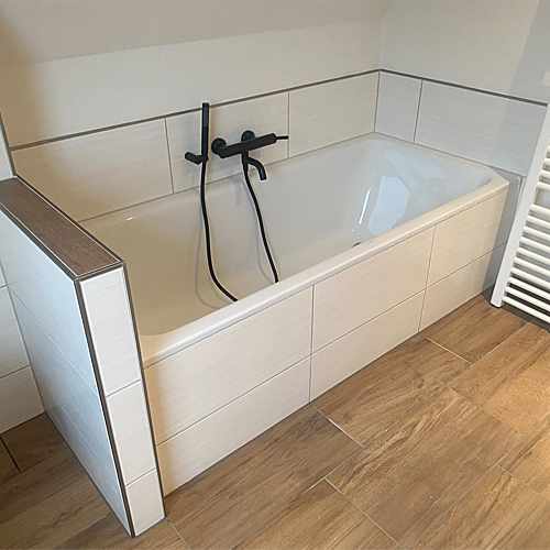 Weiß geflieste Badewanne mit schwarzer Armatur unter der Dachschräge, Boden in Holzoptik
