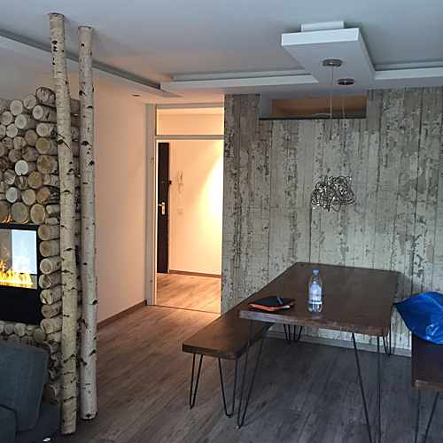 Wohnraum mit Vinylboden in Holzoptik, Birkenstämmen, Kaminofen und Esstisch vor Wand in Holzoptik